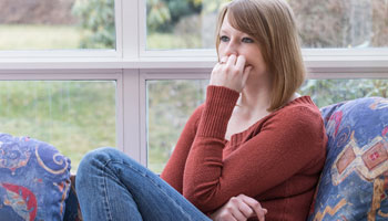 Pensive young woman dressed in a brown sweater is sitting on the sofa in conservatory and she is looking ahead